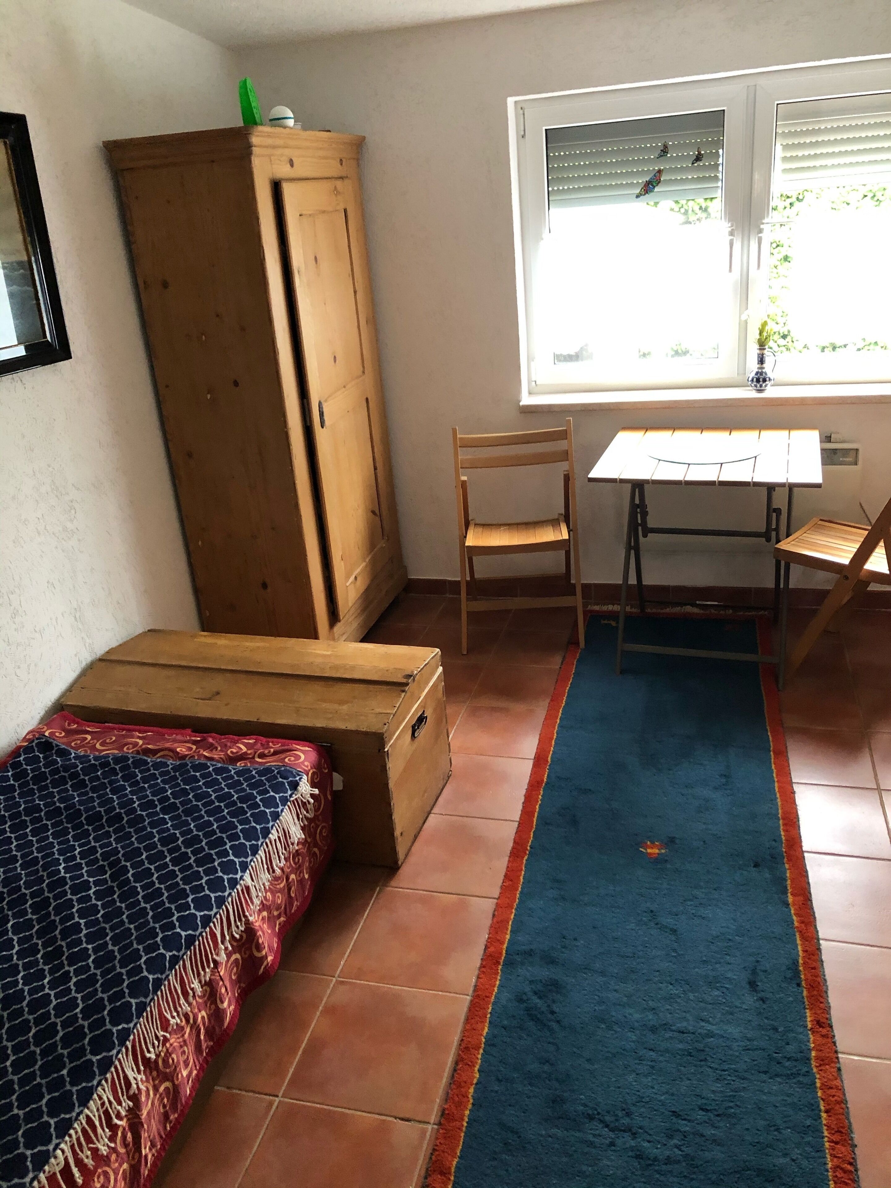 A small, furnished room with a bed, a wooden wardrobe, a desk with a chair, and a window with blinds.