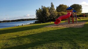 A playground with a red slide and a blue-roofed structure, located near a lake with grassy areas and trees.