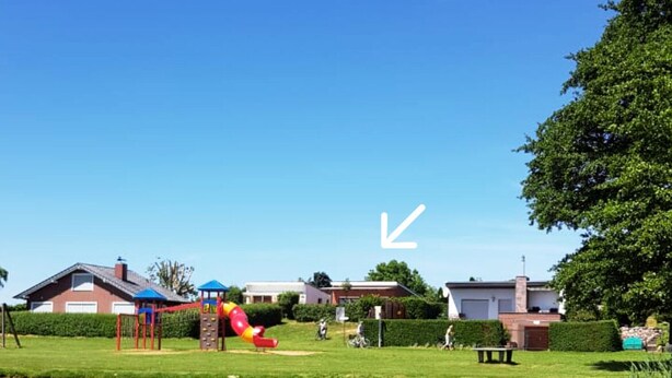 A riverside walkway with a wooden boardwalk, a playground with a red slide, and houses in the background.