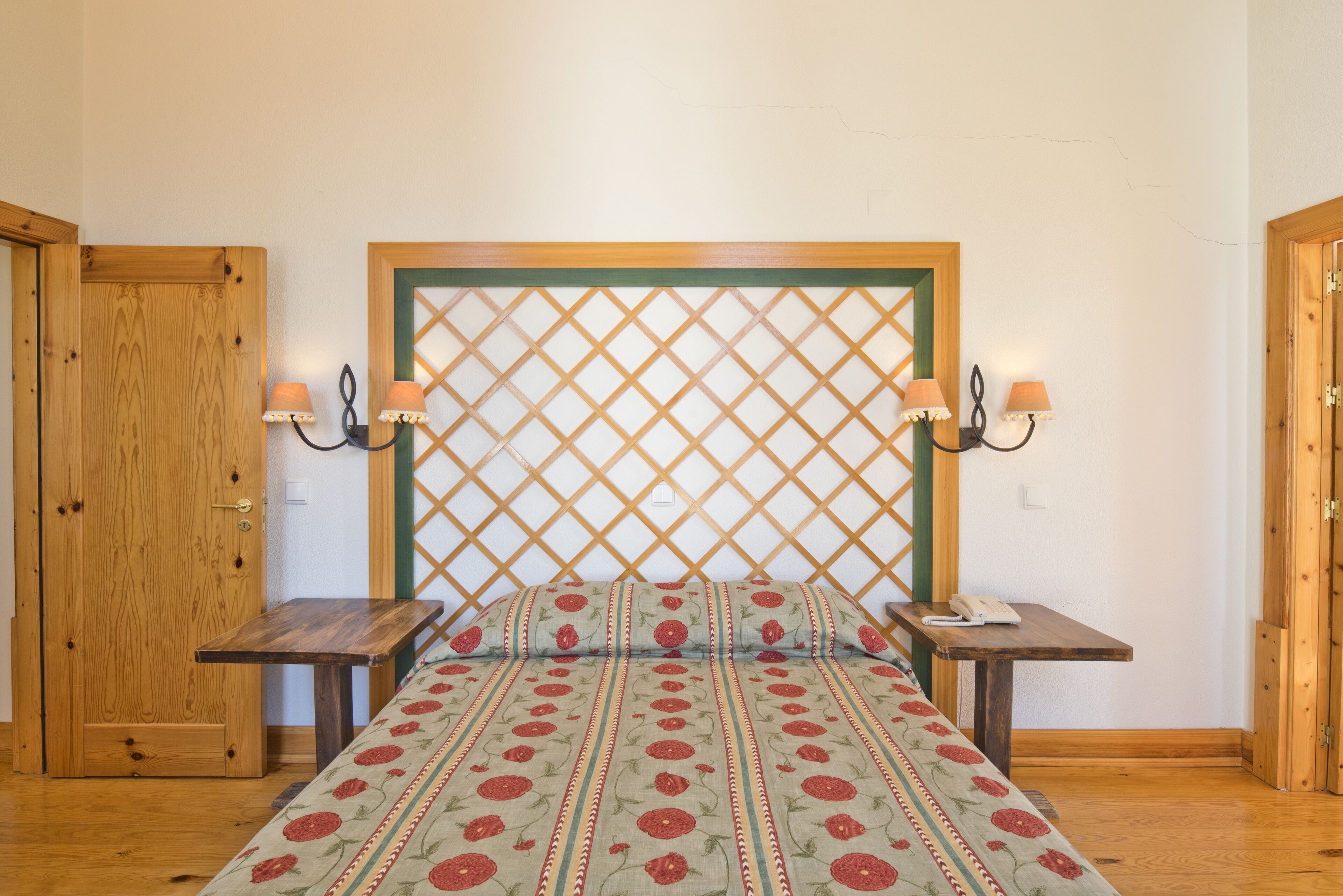 A bedroom with a wooden headboard, a bed with floral bedding, two bedside tables with lamps, and a wooden door.