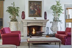 Una sala de estar con chimenea, dos sofás rojos, un otoman blanco y dos grandes macetas con plantas.