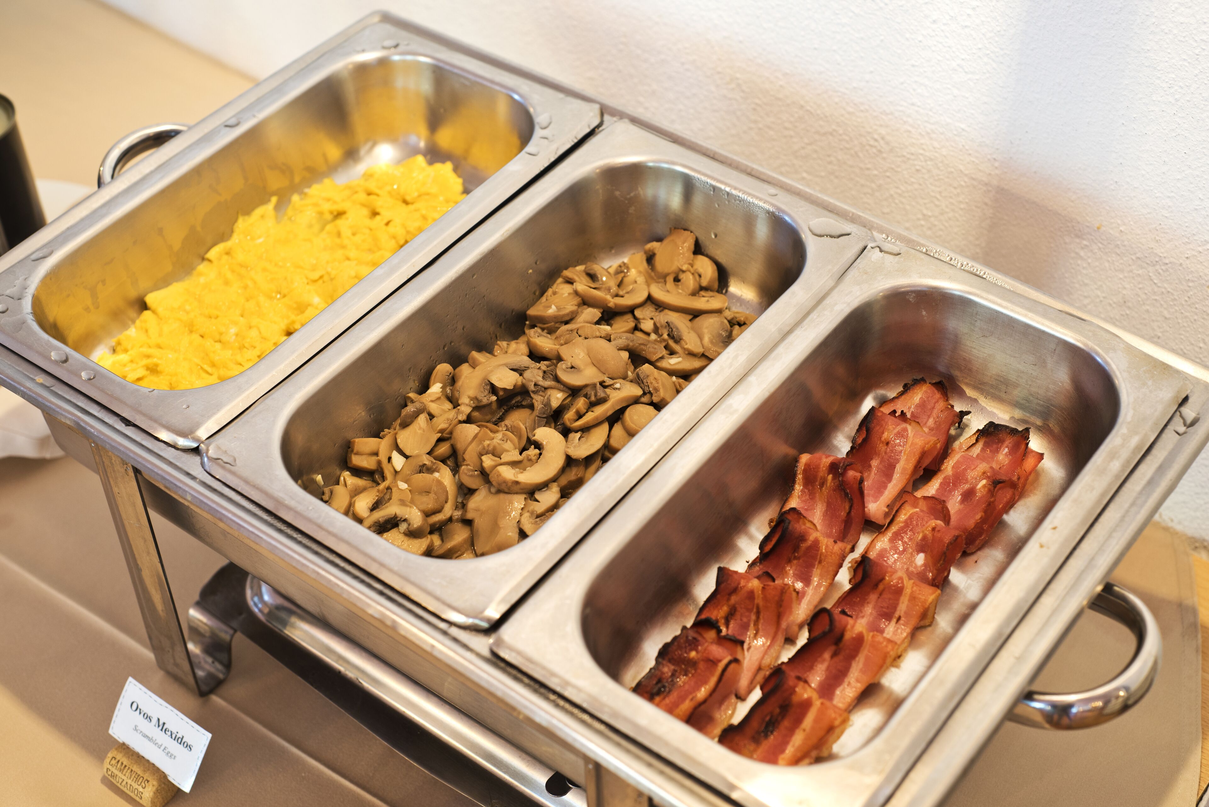 A buffet with three food stations: scrambled eggs, mushrooms, and bacon.