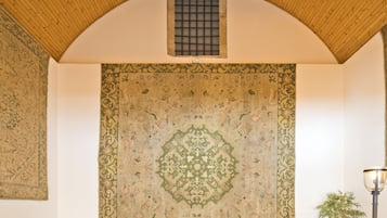 A room with a high arched wooden ceiling, a patterned tapestry on the wall, and a small window with bars.