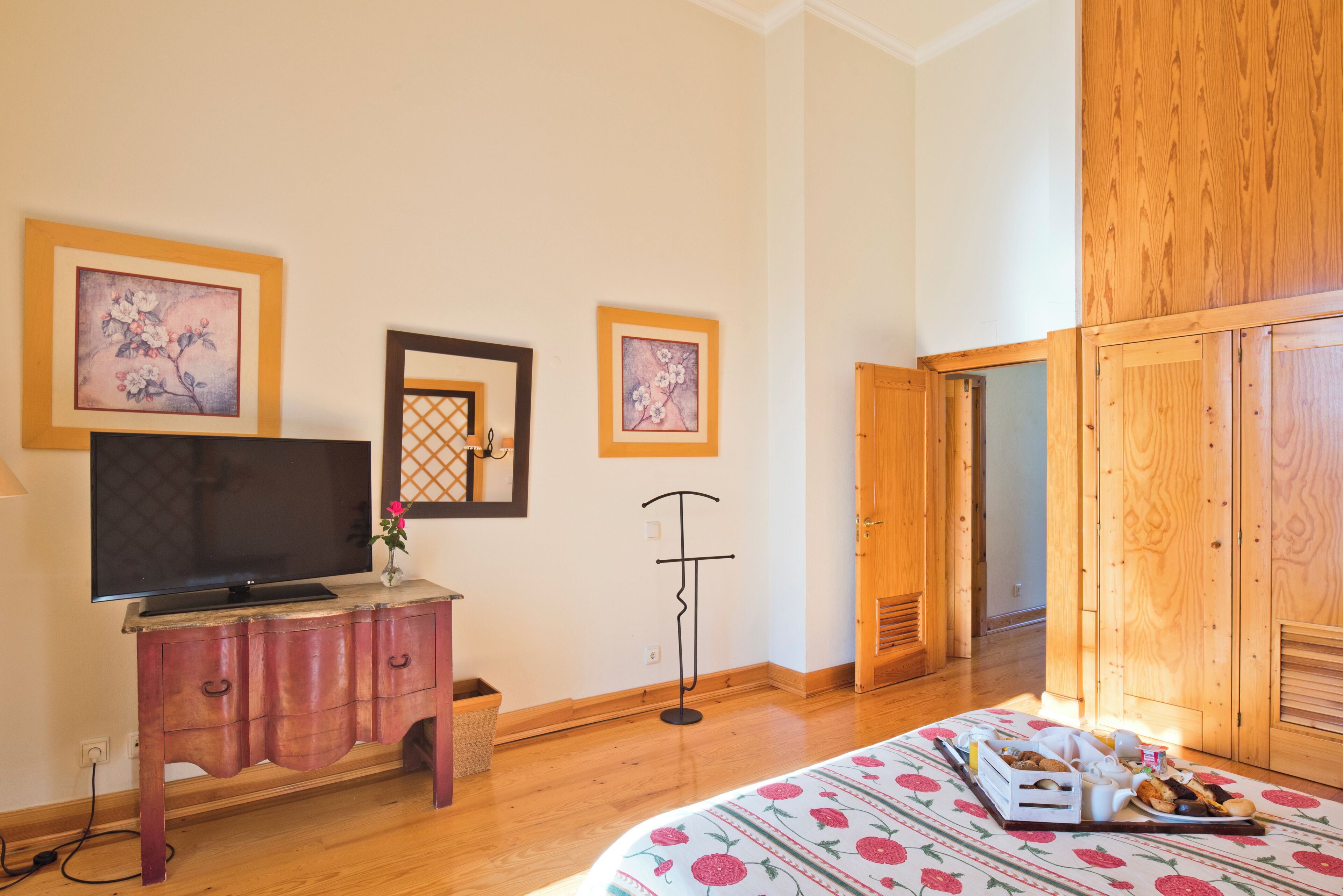 A bedroom with a bed, wooden wardrobe, a TV on a wooden cabinet, and framed floral artwork on the wall.