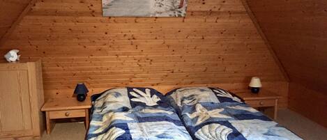 Een gezellig slaapkamer op zolder met een houten schuine plafond, een bed met een blauw-witte bedspreide, twee nachtkastjes met lampen, en een lijst met een afbeelding van een strand.