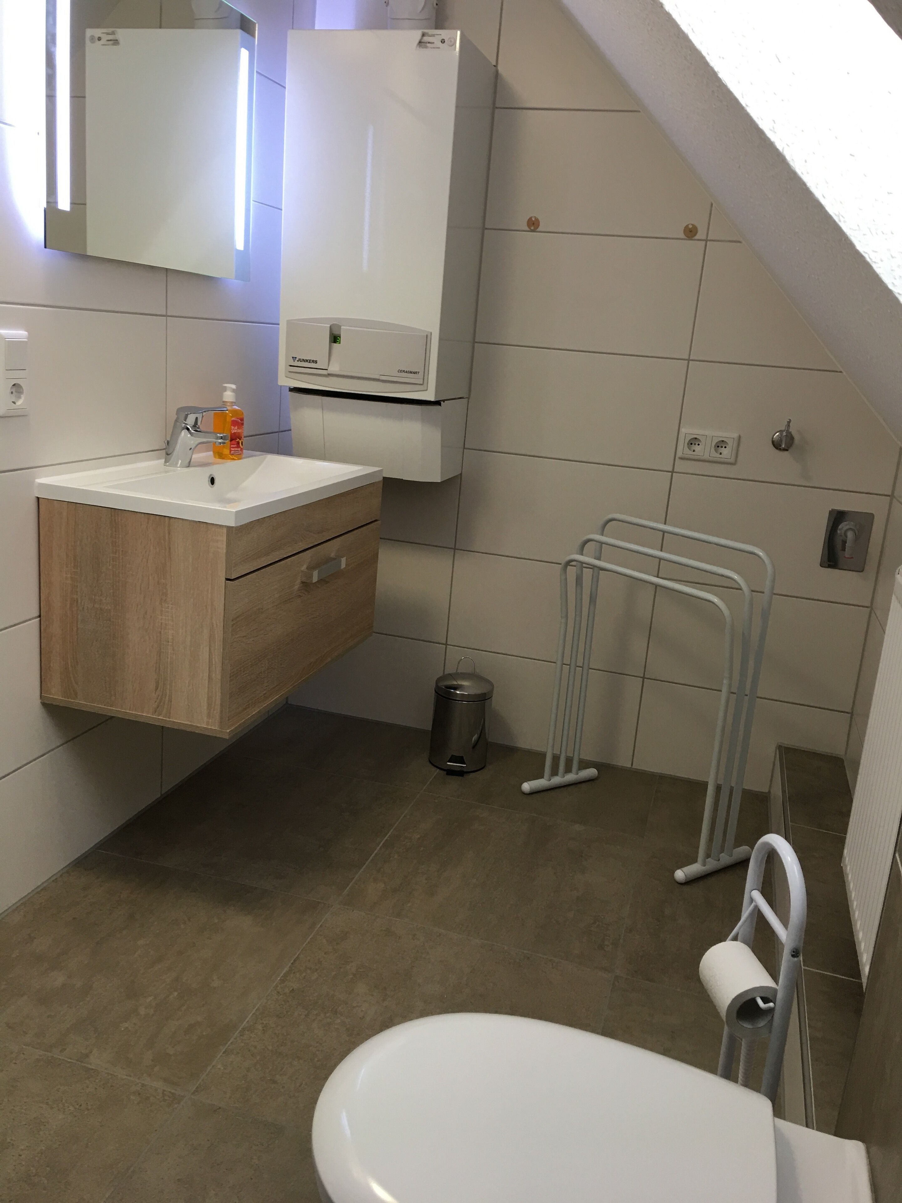 Een moderne badkamer met een houten wastafelmeubel, toilet, prullenbak en douche.