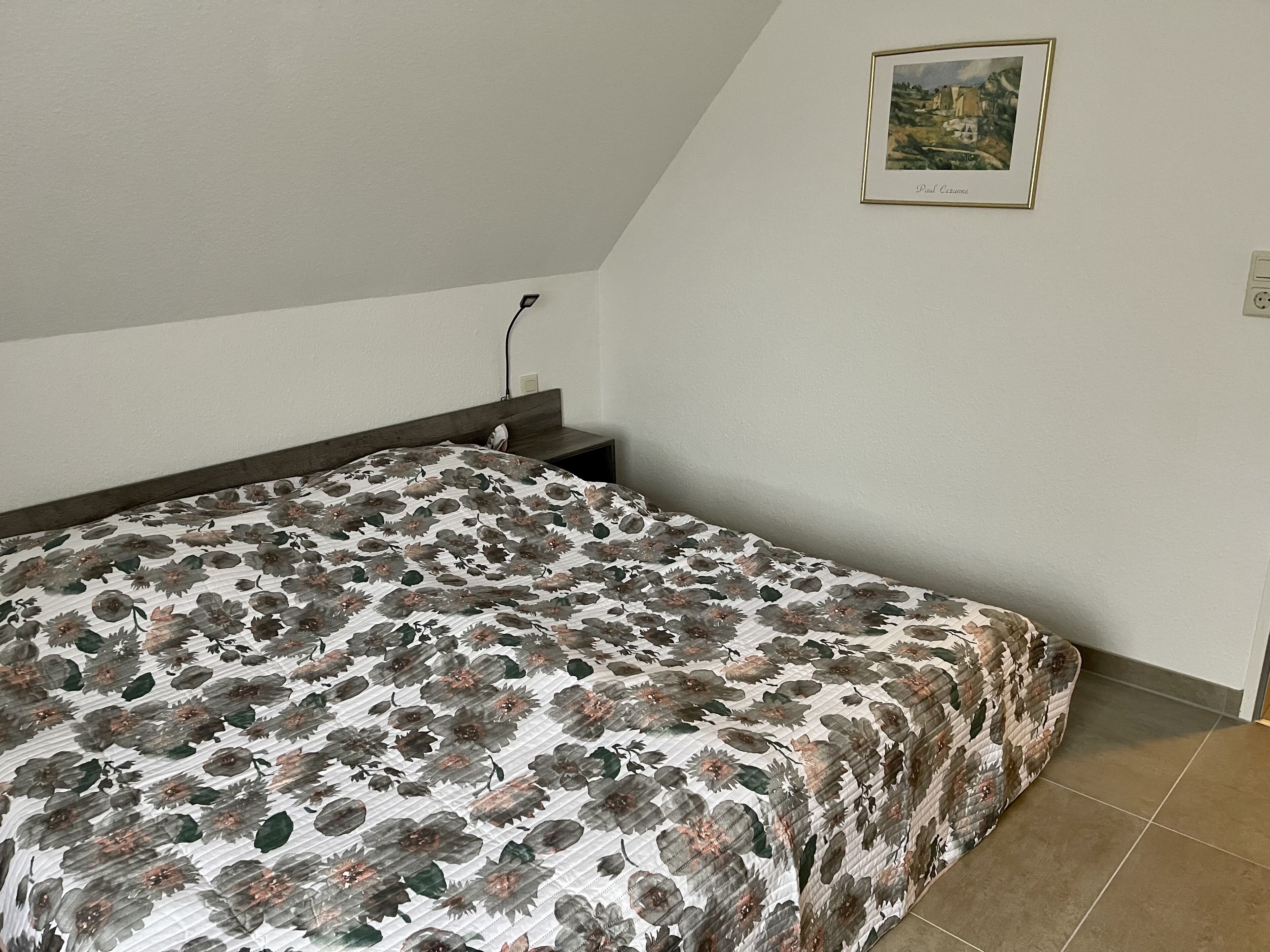 Ein Schlafzimmer mit einem Bett, einem Nachttisch, einer Wandsteckdose und einem gerahmten Bild.