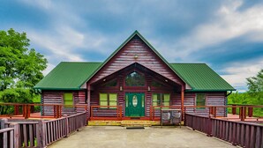 A log cabin with a green roof and a green door, surrounded by a wooden deck and railing.