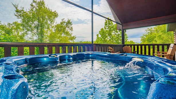 A hot tub with a wooden deck and chairs, surrounded by trees.