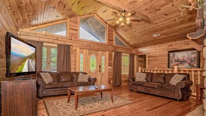 A log cabin interior with wooden floors, two leather sofas, a wooden coffee table, a TV, and a large window with a view of a road and trees.