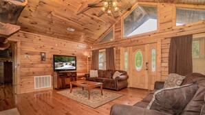 A wooden cabin interior with a wooden floor, a brown leather sofa, a television, and a ceiling fan.