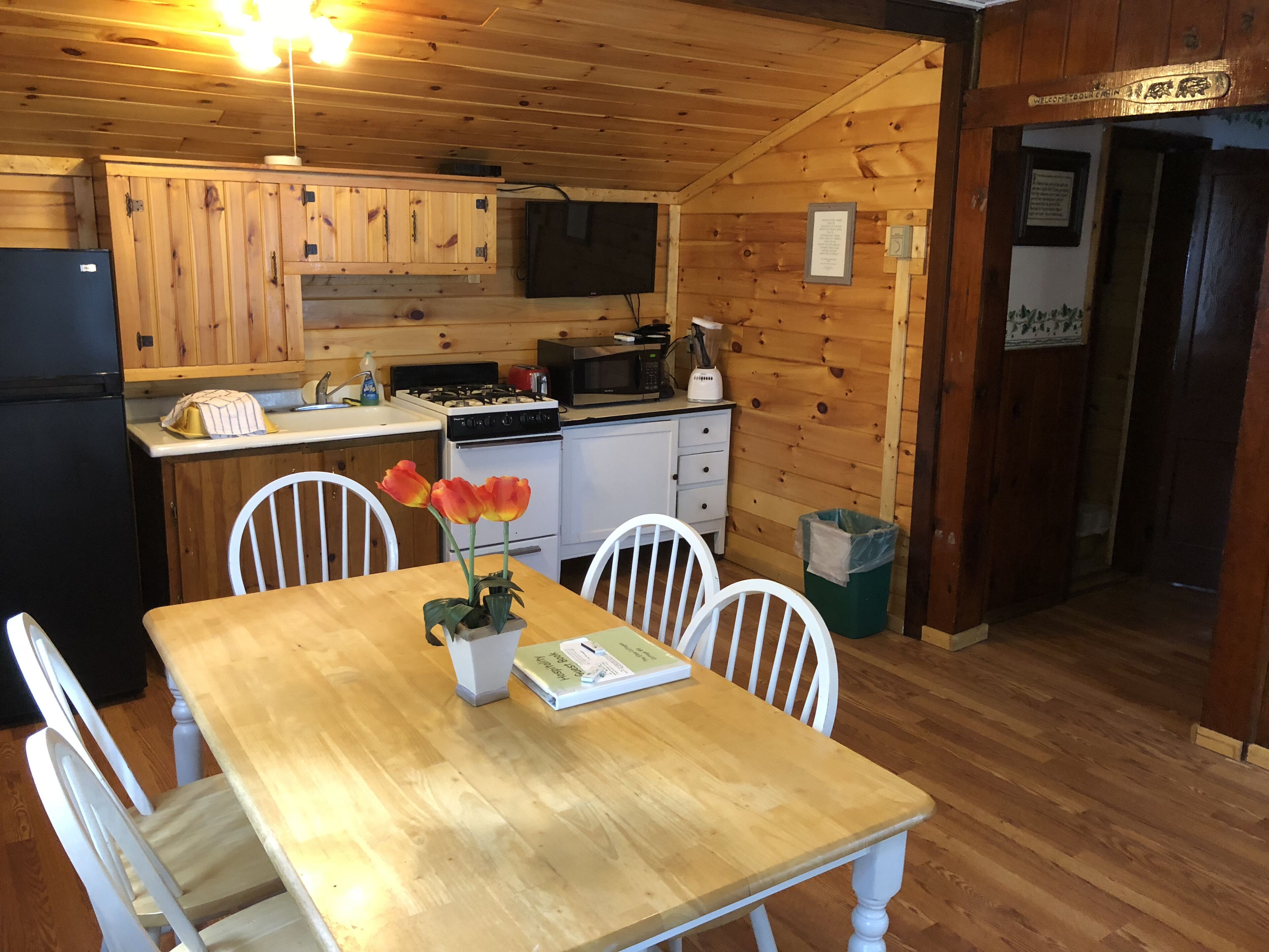 A wooden cabin-style kitchen with a dining table, chairs, a refrigerator, stove, microwave, and a trash can.