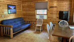 Ein Holzblockhaus-Innenraum mit einer Couch, einem Esstisch, Stühlen, einem Fernseher und einem Fenster mit Jalousien.