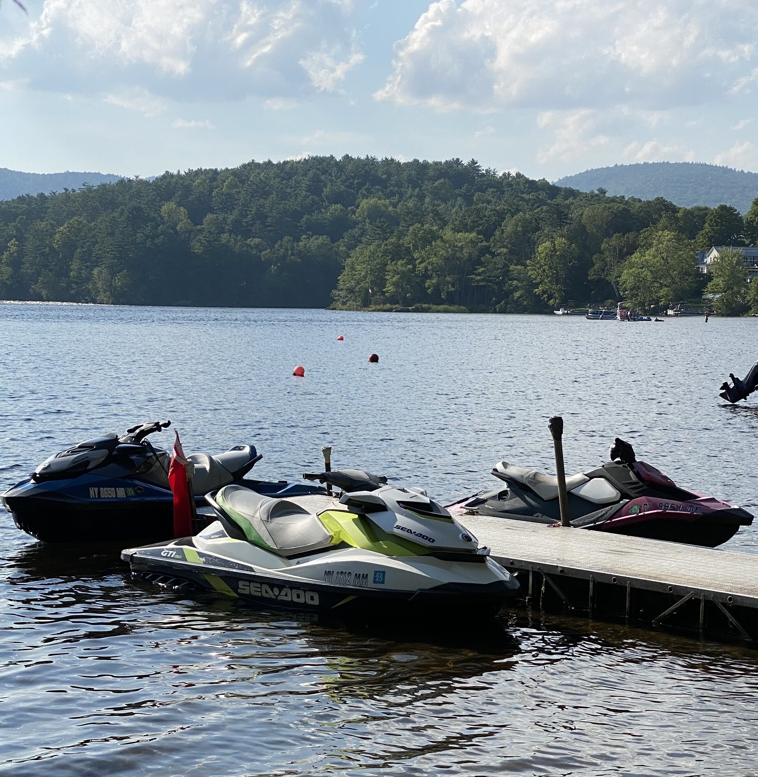 A dock with several jet skis and a boat, surrounded by calm water and a forested hillside.