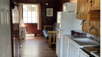 A narrow, wooden-paneled kitchen with a refrigerator, sink, and wooden cabinets. A window with curtains allows natural light to enter.