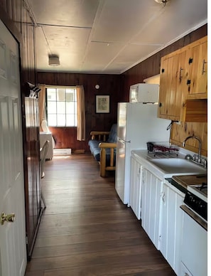 Una cocina estrecha con paneles de madera, que incluye un refrigerador, un lavamanos y armarios de madera. Una ventana con cortinas deja entrar luz natural.