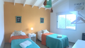 A room with two beds, each with a teal bedspread and white pillows, a small bedside table with a lamp, and a window with a view of trees.