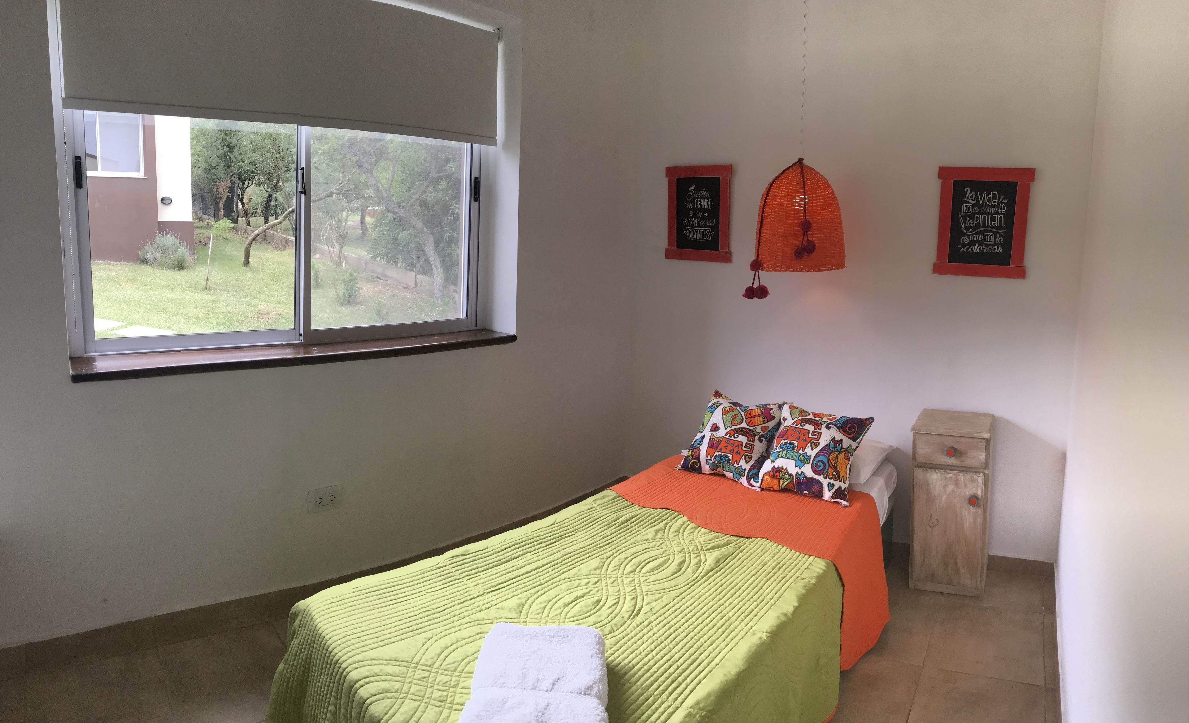 A bedroom with a bed, a window, a wall-mounted shelf, and a small cabinet.