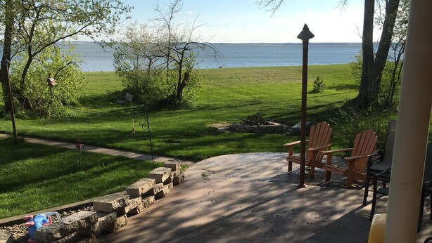 A covered patio with a view of a grassy area and a body of water. There are wooden chairs and a table, and a fire pit is visible.