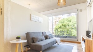 A modern living room with a grey sofa, a small side table with a plant, a large window with a view of greenery and buildings, and a wooden floor. - Fitz Roy by Be Local Argentina (Buenos Aires)