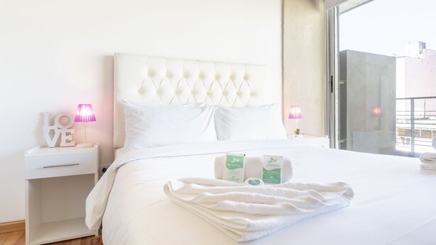 A neatly made bed with white linens, a tufted headboard, and a view of the city through a large window. - Fitz Roy by Be Local Argentina (Buenos Aires)