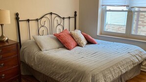 A bedroom with a bed, a nightstand, a lamp, a window with blinds, and a patterned rug. - 50's Classic: Cozy, convenient, comfortable (St Louis)