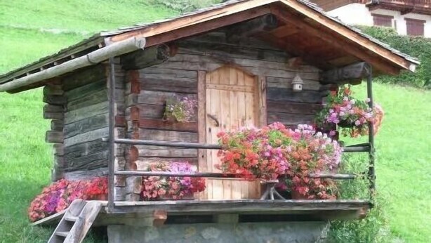 Una cabaña rústica de troncos con puerta de madera, un pequeño porche con arreglos florales y una escalera de madera.