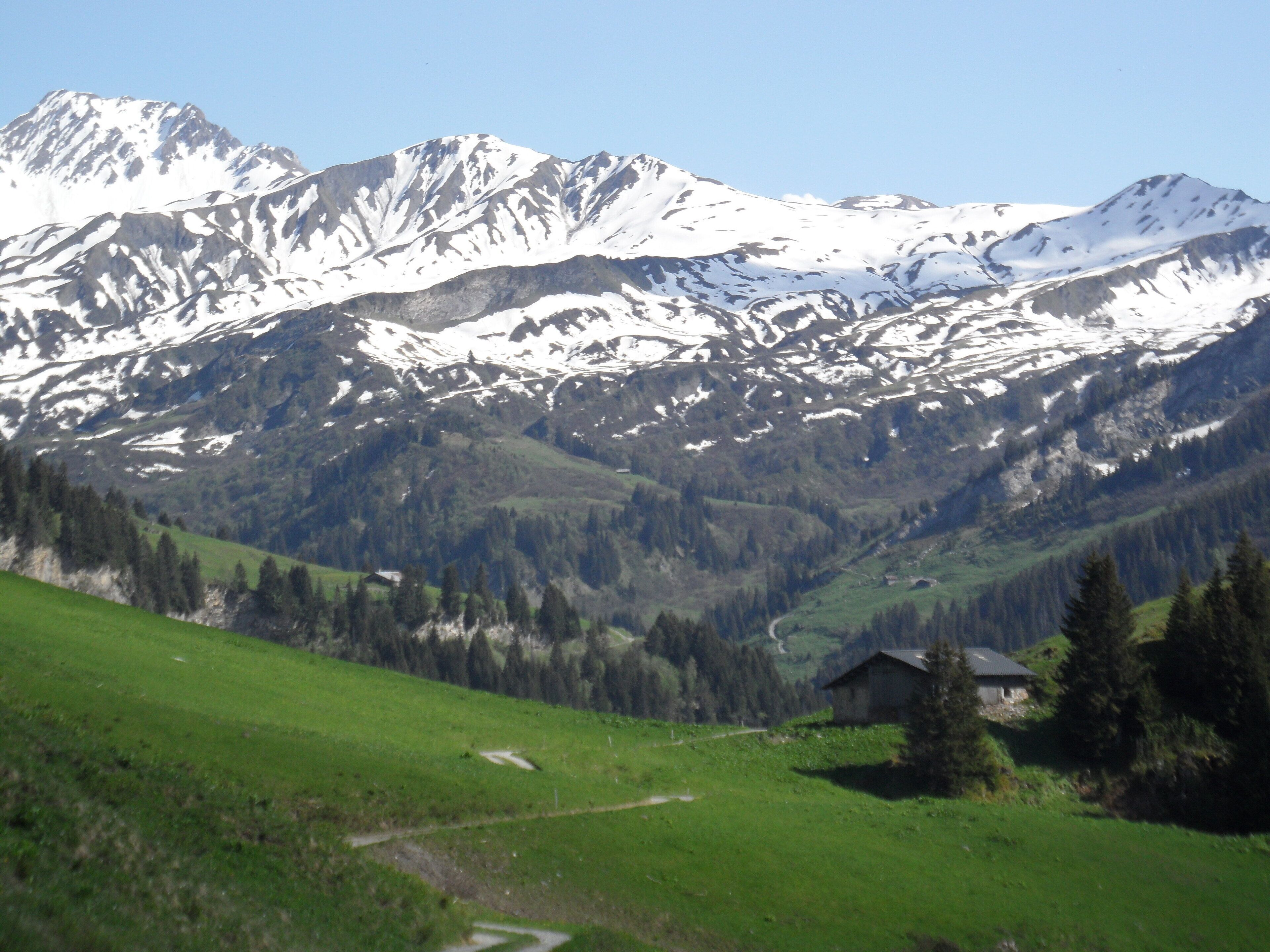 Een bergachtig landschap met besneeuwde toppen, een groene vallei en een klein huis op de voorgrond.