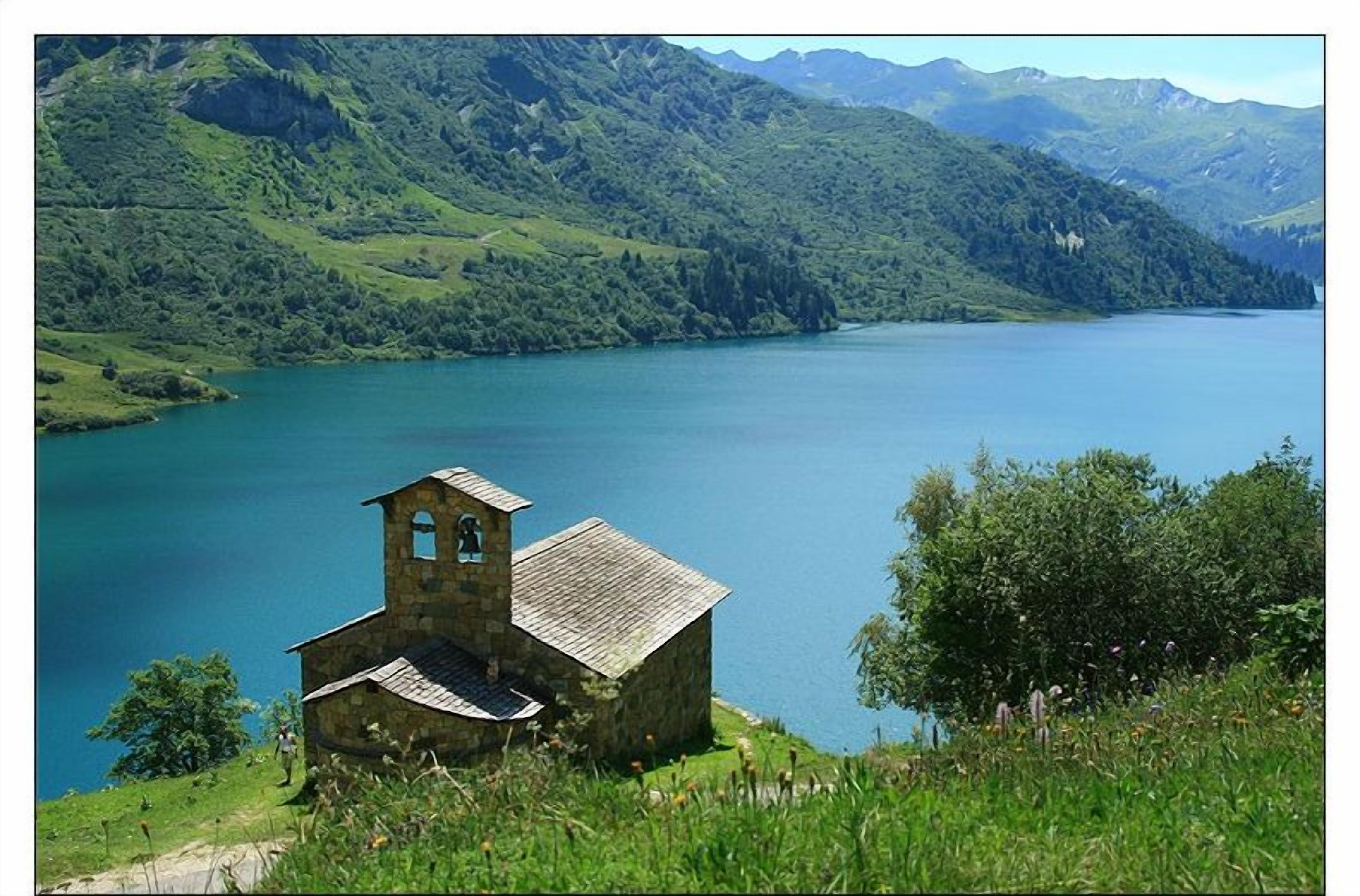 A small stone chapel with a bell tower, surrounded by lush green hills and a calm lake.