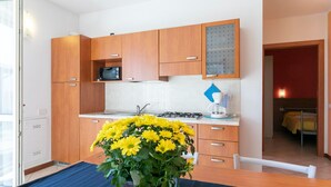 A modern kitchen with wooden cabinets, a gas stove, and a sink. There is a blue mat on the floor and a potted plant with yellow flowers on the table. - Lake Como - Residence La Collina - Pera (Vercana)