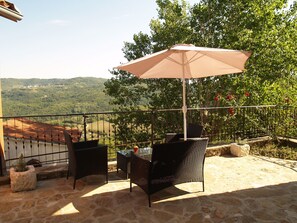 Un patio con suelo de piedra, dos sillas de mimbre, una mesa y un gran paraguas color beige.