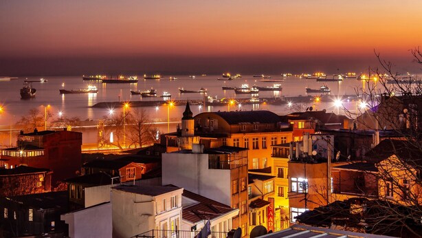 Un paisaje urbano costero al anochecer, con embarcaciones e edificios iluminados.
