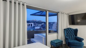 Une chambre d’hôtel moderne avec une grande fenêtre, un lit, un fauteuil bleu et un petit pouf.