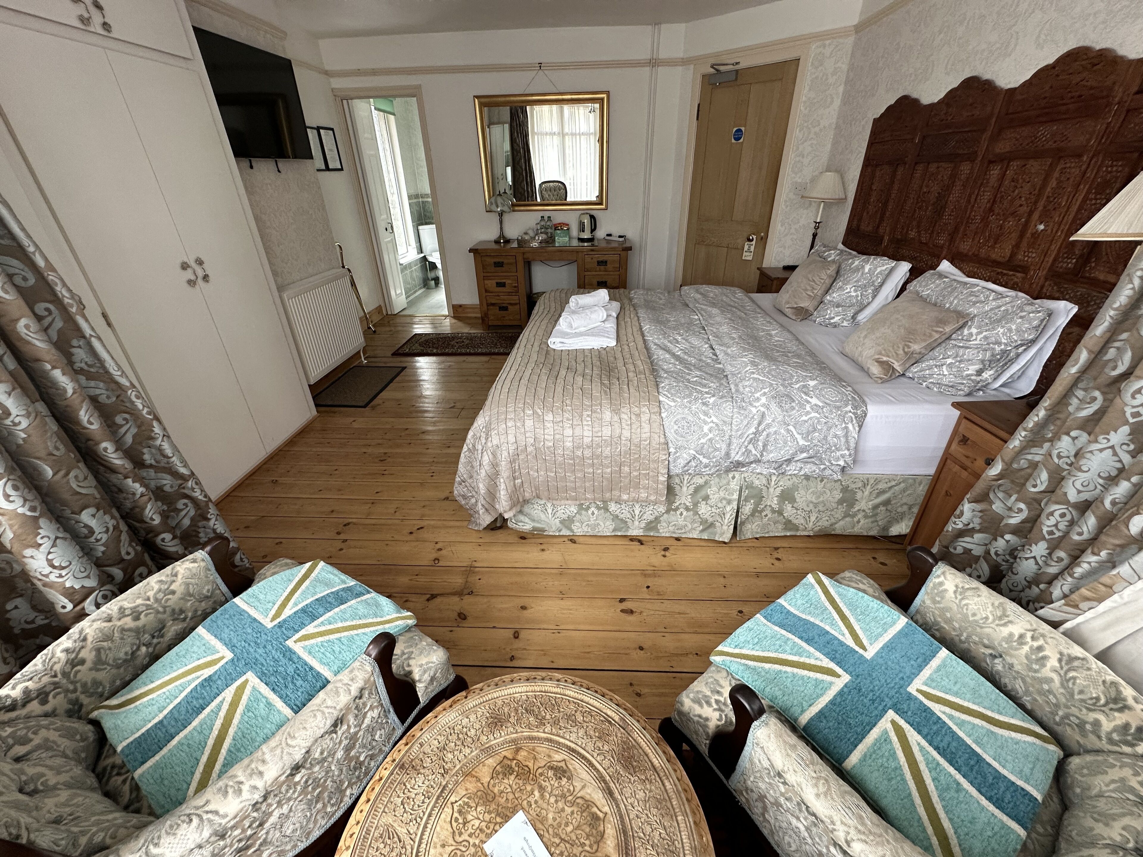 A spacious bedroom with a large bed, wooden floor, and a small sitting area with patterned cushions.