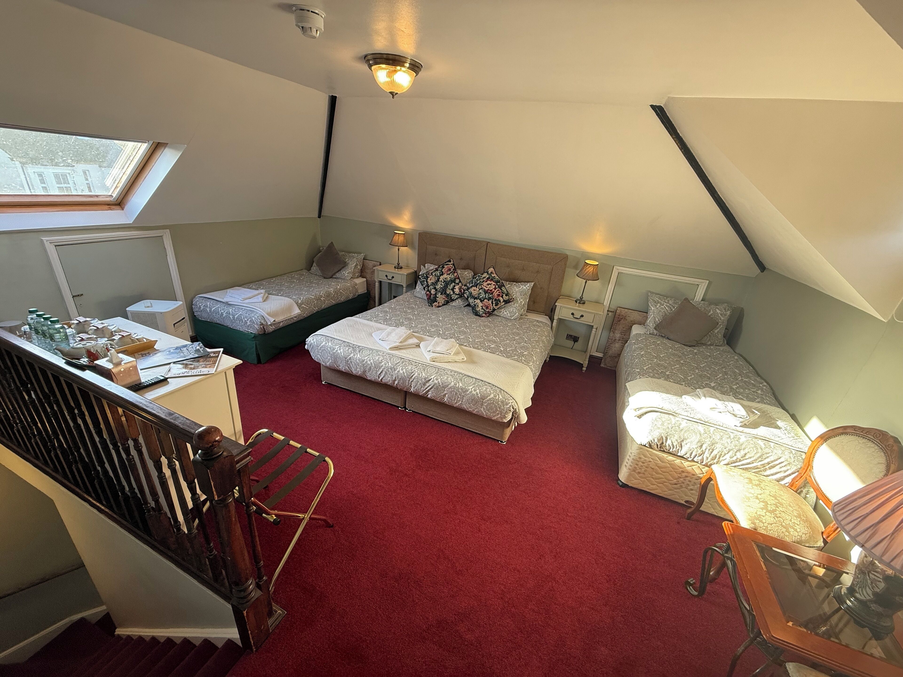 Une chambre d’hôtel avec deux lits, un bureau, une chaise, une moquette rouge et une lucarne.
