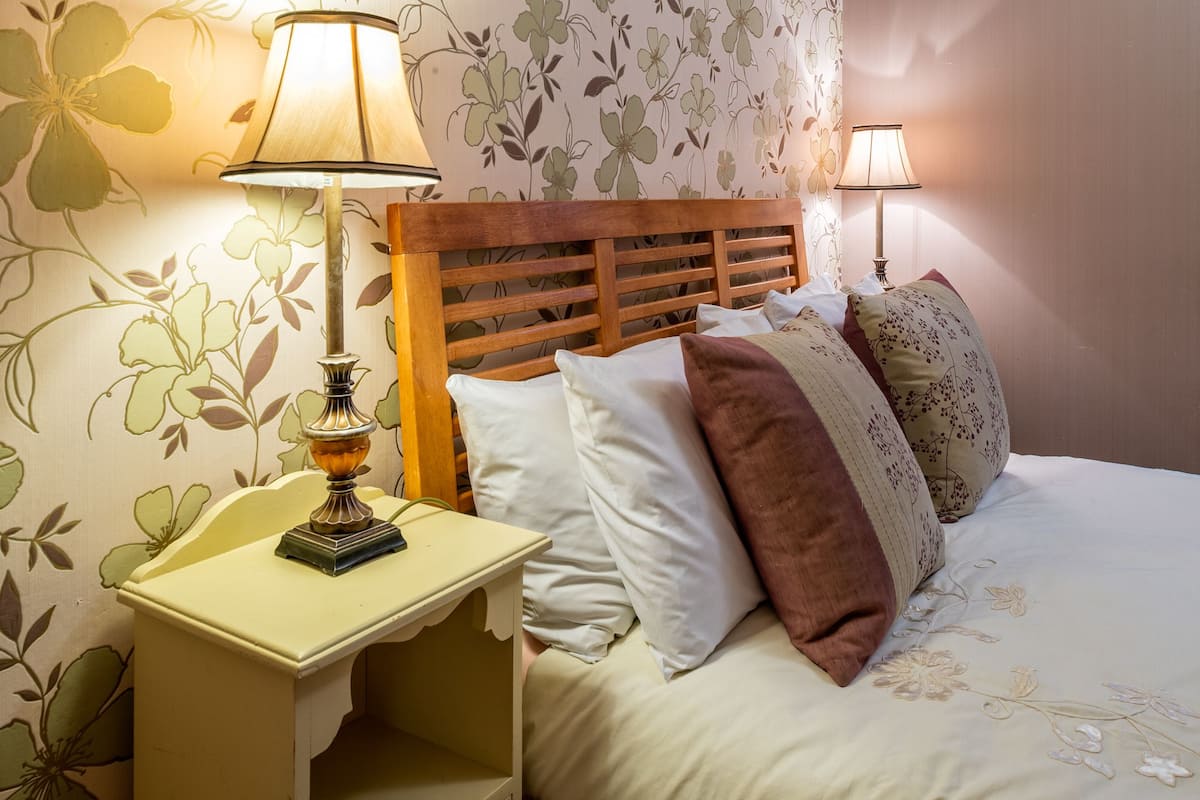 A neatly made bed with a wooden headboard, a bedside table with a lamp, and floral wallpaper.
