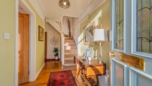 Ein Flur mit Holzboden, einem roten Teppich mit Muster, einer Treppe, einer Uhr, einer Vase mit Zweigen, einem Tisch mit Lampe und einem Fenster mit dekorativem Gitter.