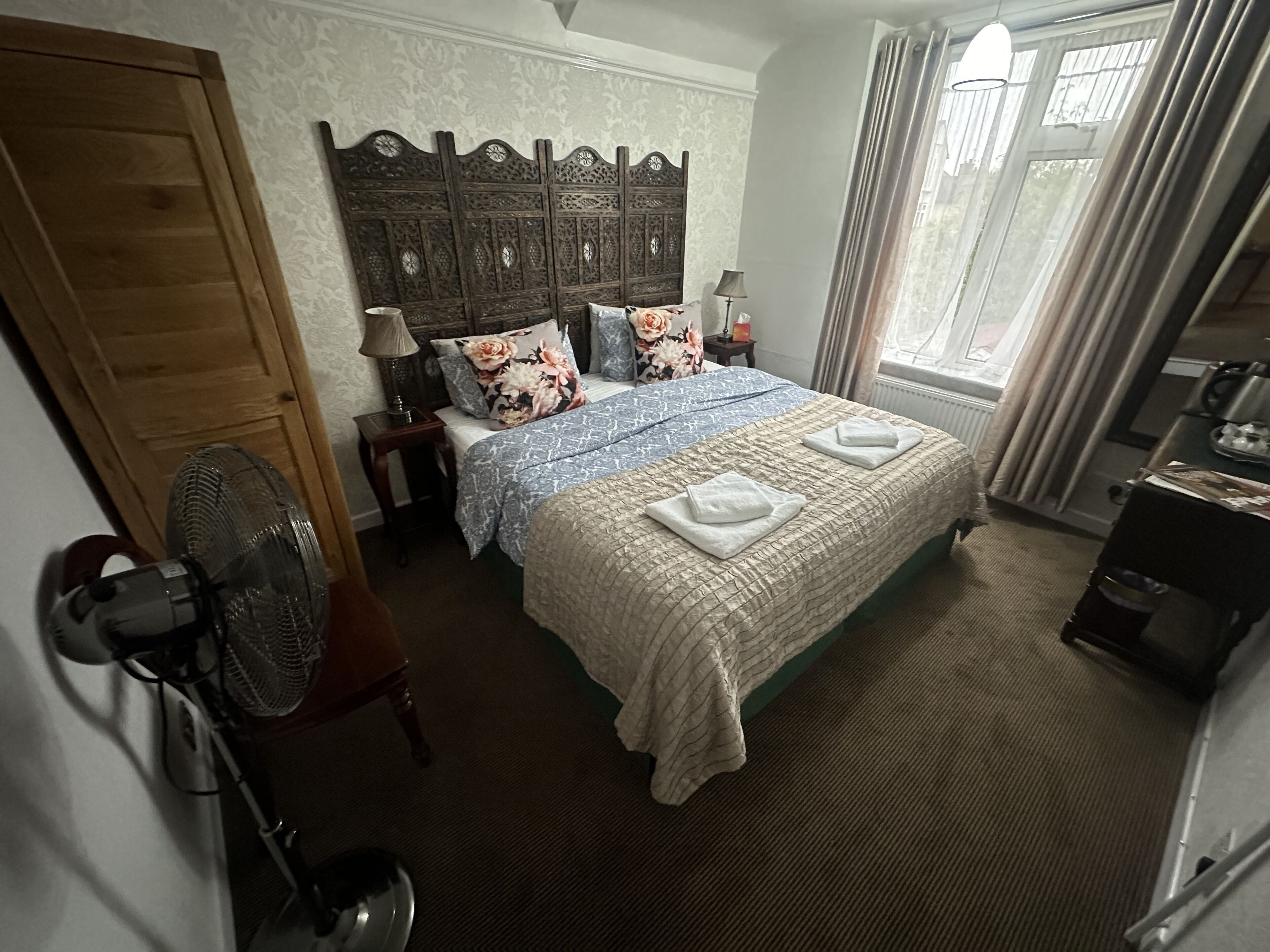 Een slaapkamer met een groot bed, een houten hoofdeinde, een nachtkastje, een lamp, een ventilator en een raam met gordijnen.