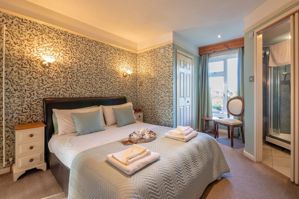 A bedroom with a large bed, two bedside tables, a chair, and a bathroom with a shower and a mirror.