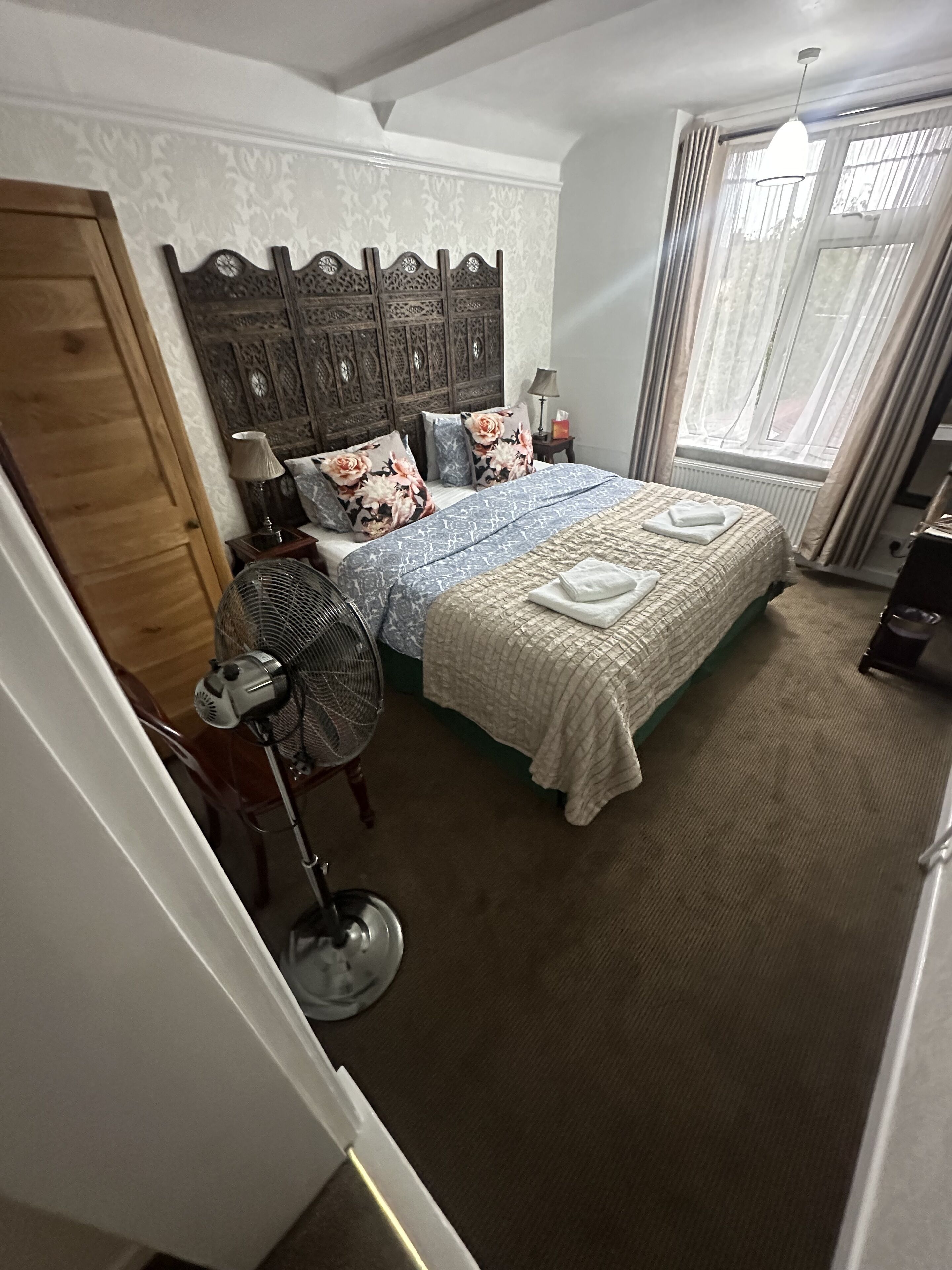 Une chambre avec un grand lit, une tête de lit en bois, un ventilateur sur pied, un radiateur et une fenêtre avec des rideaux.