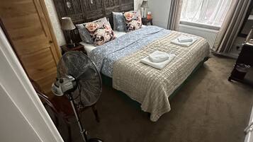 Une chambre avec un grand lit, une tête de lit en bois, un ventilateur sur pied, un radiateur et une fenêtre avec des rideaux.