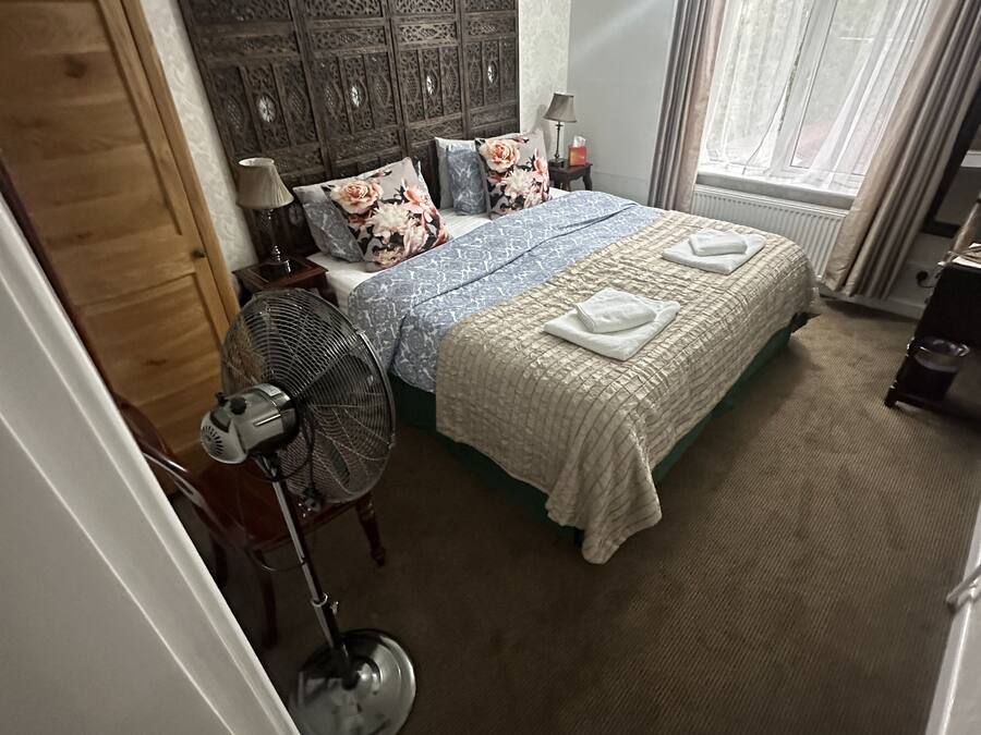 Une chambre avec un grand lit, une tête de lit en bois, un ventilateur sur pied, un radiateur et une fenêtre avec des rideaux.