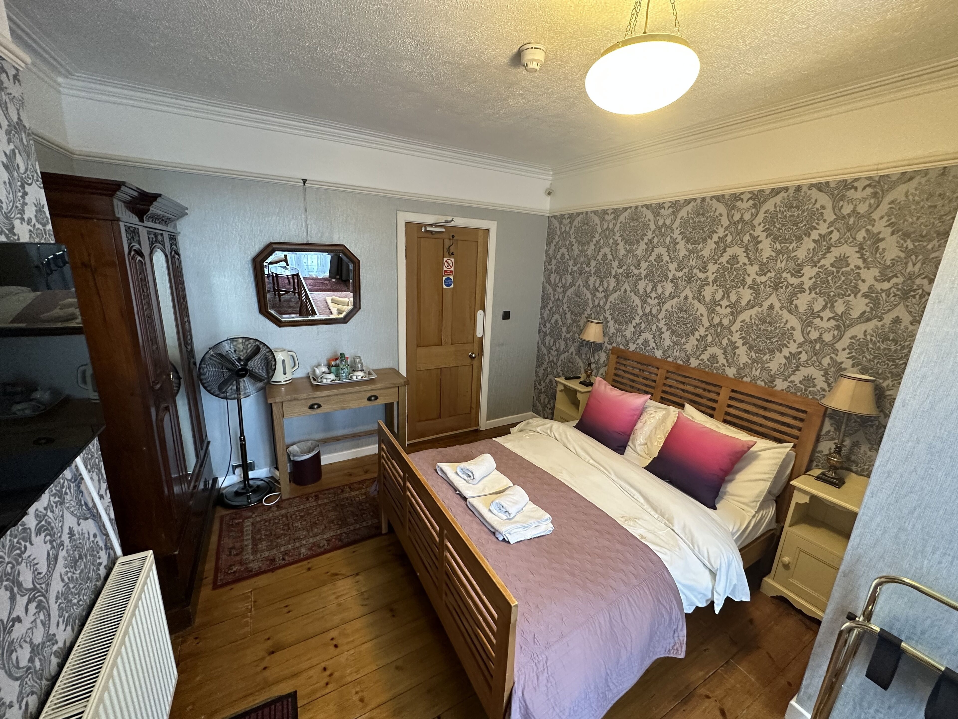 A bedroom with a wooden bed, a nightstand, a desk with a mirror, a fan, and a wardrobe.