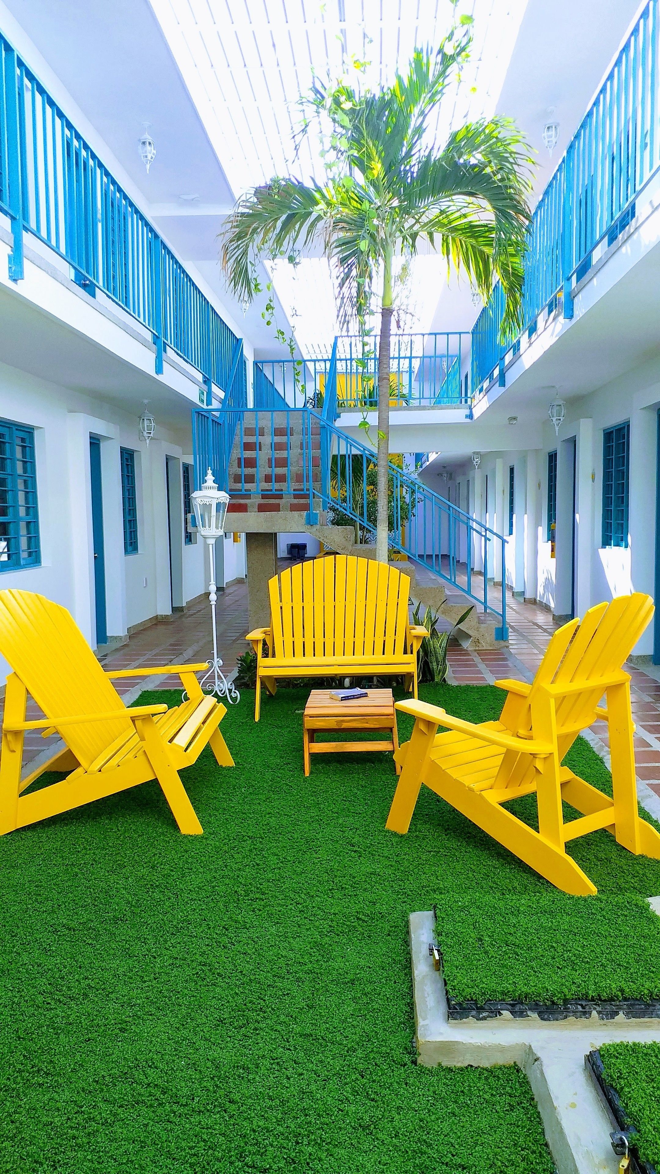 A courtyard with artificial grass, yellow Adirondack chairs, a wooden bench, and a palm tree.