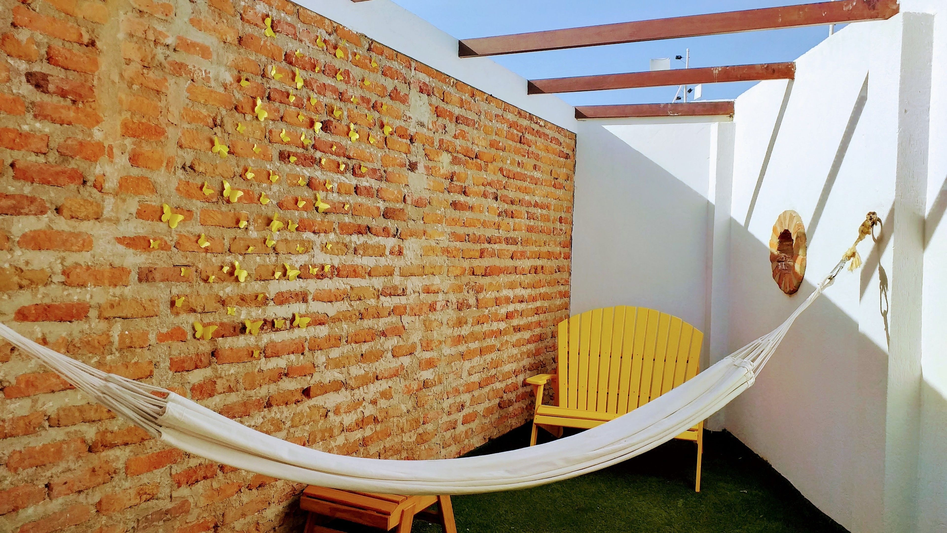 Una hamaca atada entre un muro de ladrillo y una pared blanca, con una silla amarilla y un taburete de madera cerca.