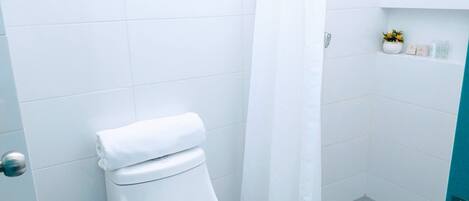 Un baño pequeño y blanco con inodoro, cortina de ducha y estante con artículos de aseo.