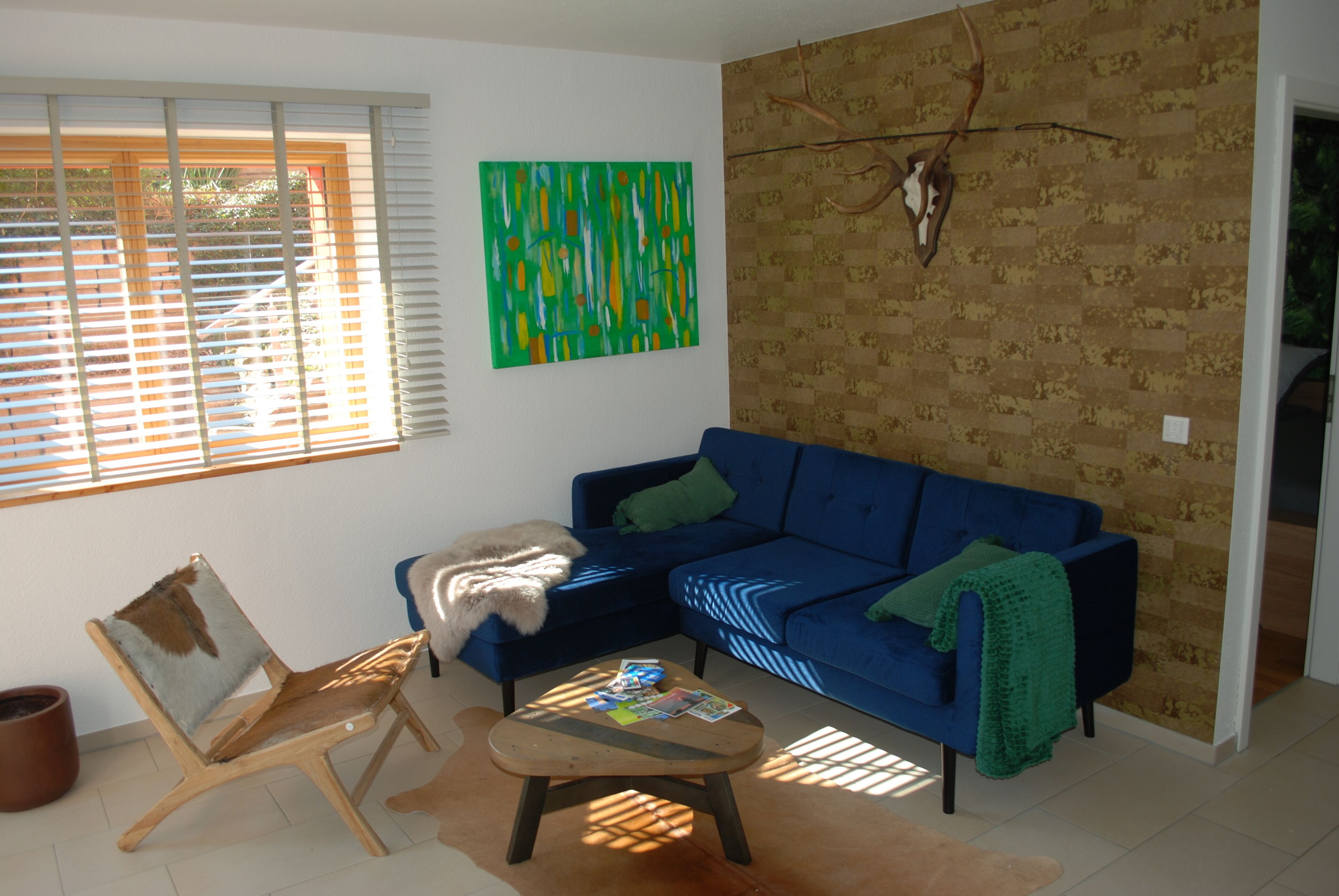 A living room with a blue sofa, a wooden chair, a coffee table with magazines, a green blanket, and a wall-mounted deer skull.