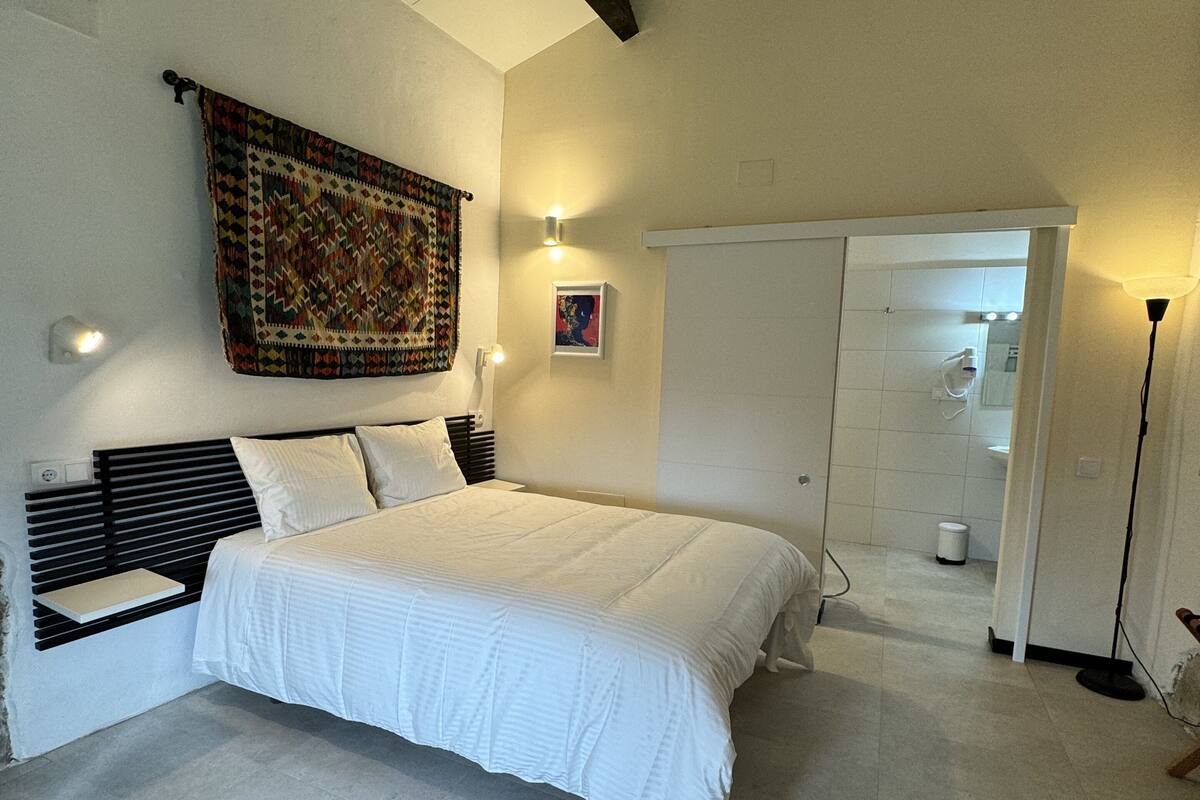 Un dormitorio con una cama grande, un cabecero, un tapiz, una lámpara de pared y una puerta que da a un baño.