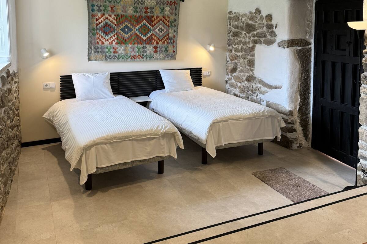 Una habitación con dos camas, una pared de piedra, una puerta negra y un colcha colgada.