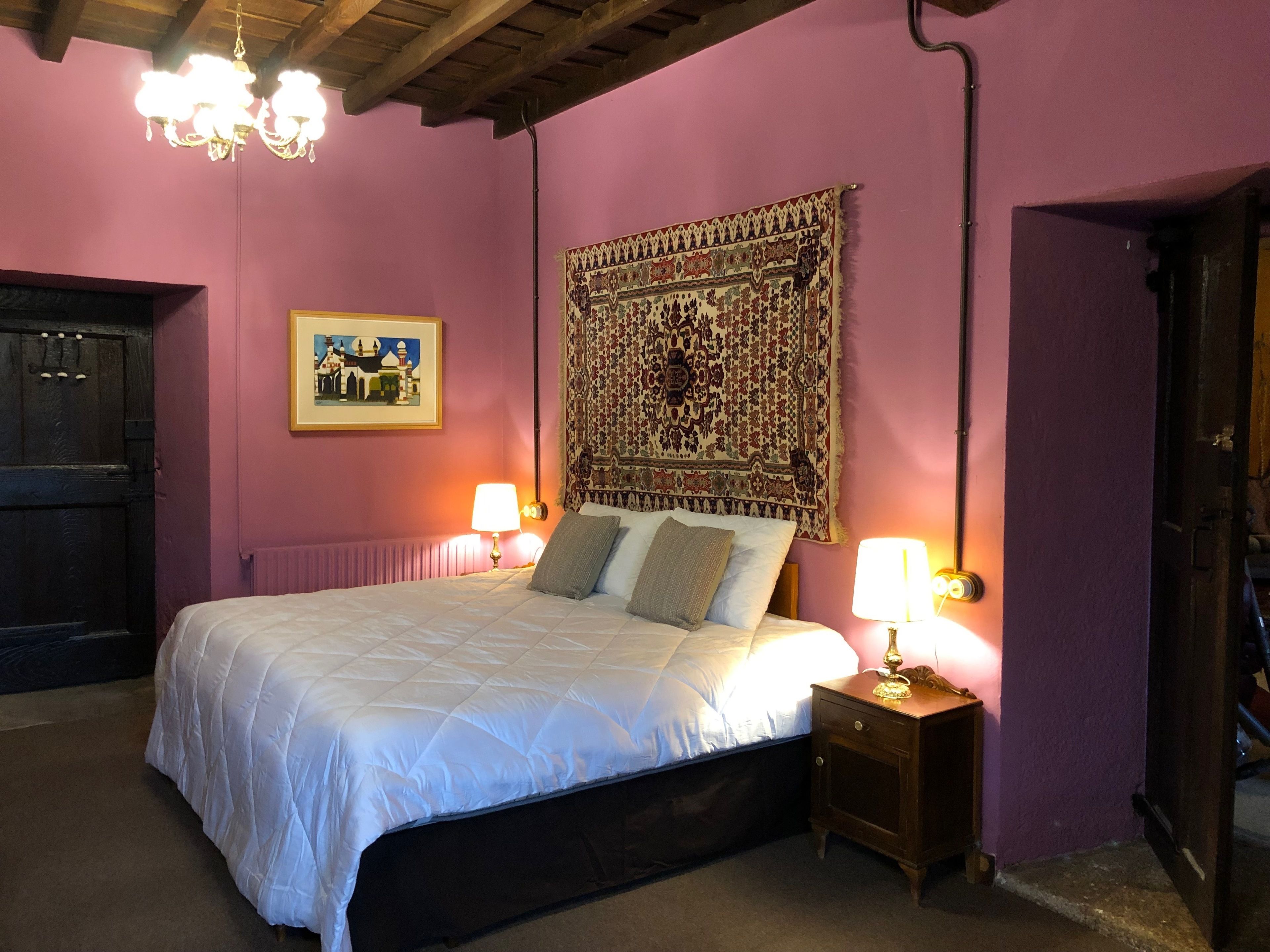 A bedroom with a large bed, two bedside lamps, a tapestry, and a door.